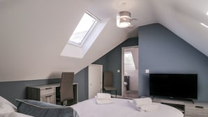A modern bedroom with a skylight, a bed, a desk, a chair, a television, and a bathroom visible through an open door. - Magnolia House (Reading)