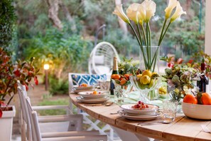 Un espace de restauration en plein air avec une table en bois dressée pour un repas, entourée de verdure et agrémentée d’une chaise blanche.