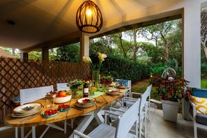 Un espace de restauration en plein air avec une table en bois dressée pour un repas, entourée de chaises blanches, et un luminaire décoratif suspendu au-dessus.