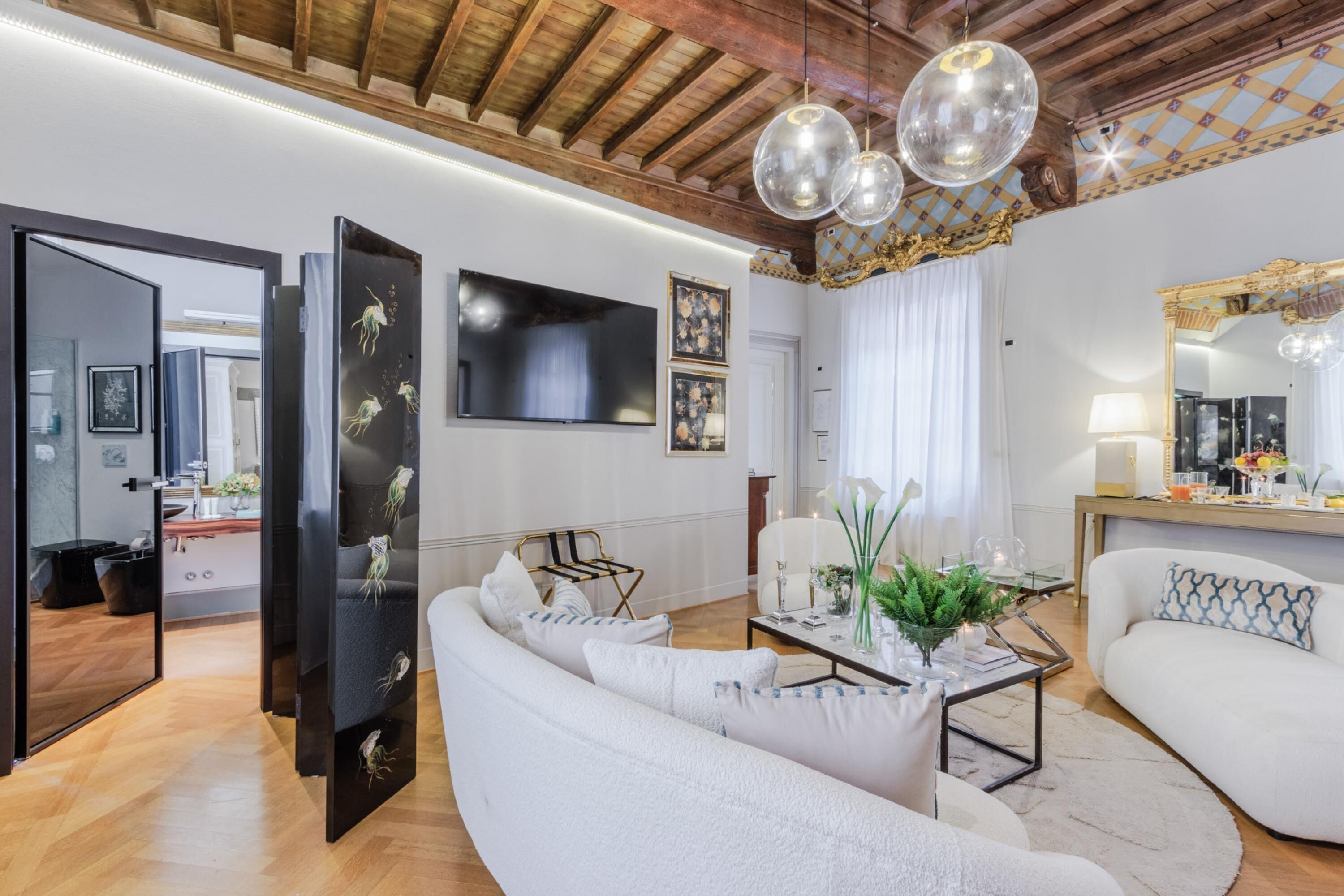 Un salón moderno con un sofá blanco, una mesa de centro de cristal y un televisor de pantalla plana grande. El techo es de madera con vigas a la vista y un candelabro.