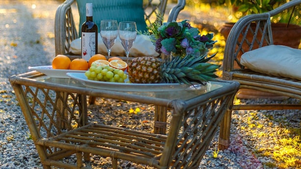 A wicker table with a bottle, glasses, fruits, and a bouquet of flowers on a gravel path. - Amore Farmhouse Retreat (Lucca)