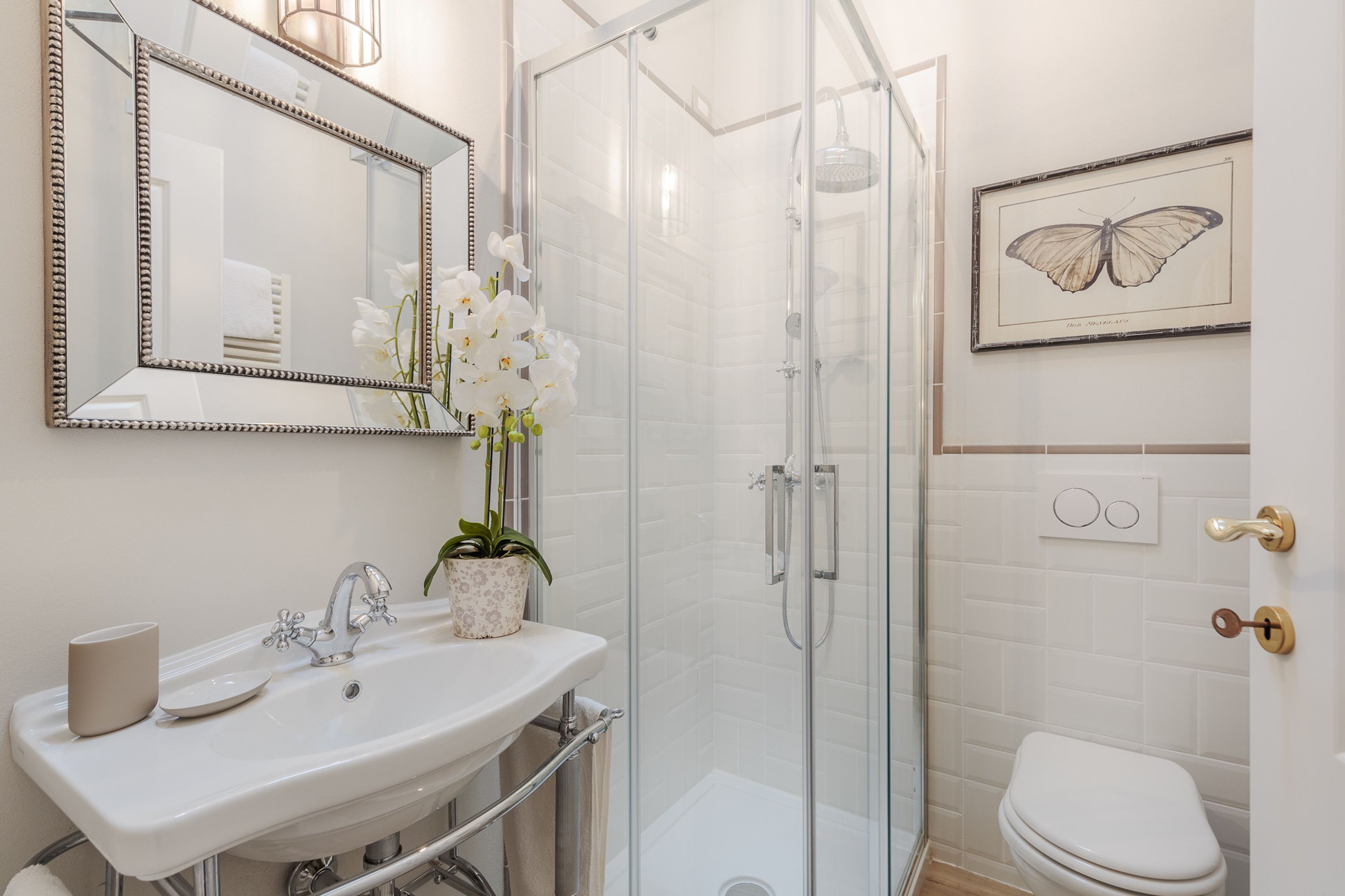 A bathroom with a sink, mirror, shower, toilet, and a framed butterfly artwork on the wall.