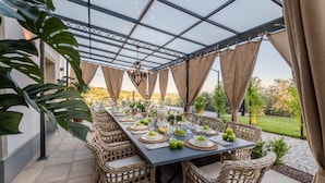 A covered outdoor dining area with a long table set for a meal, surrounded by wicker chairs. - Villa Asti in Segromigno in Monte (Capannori)