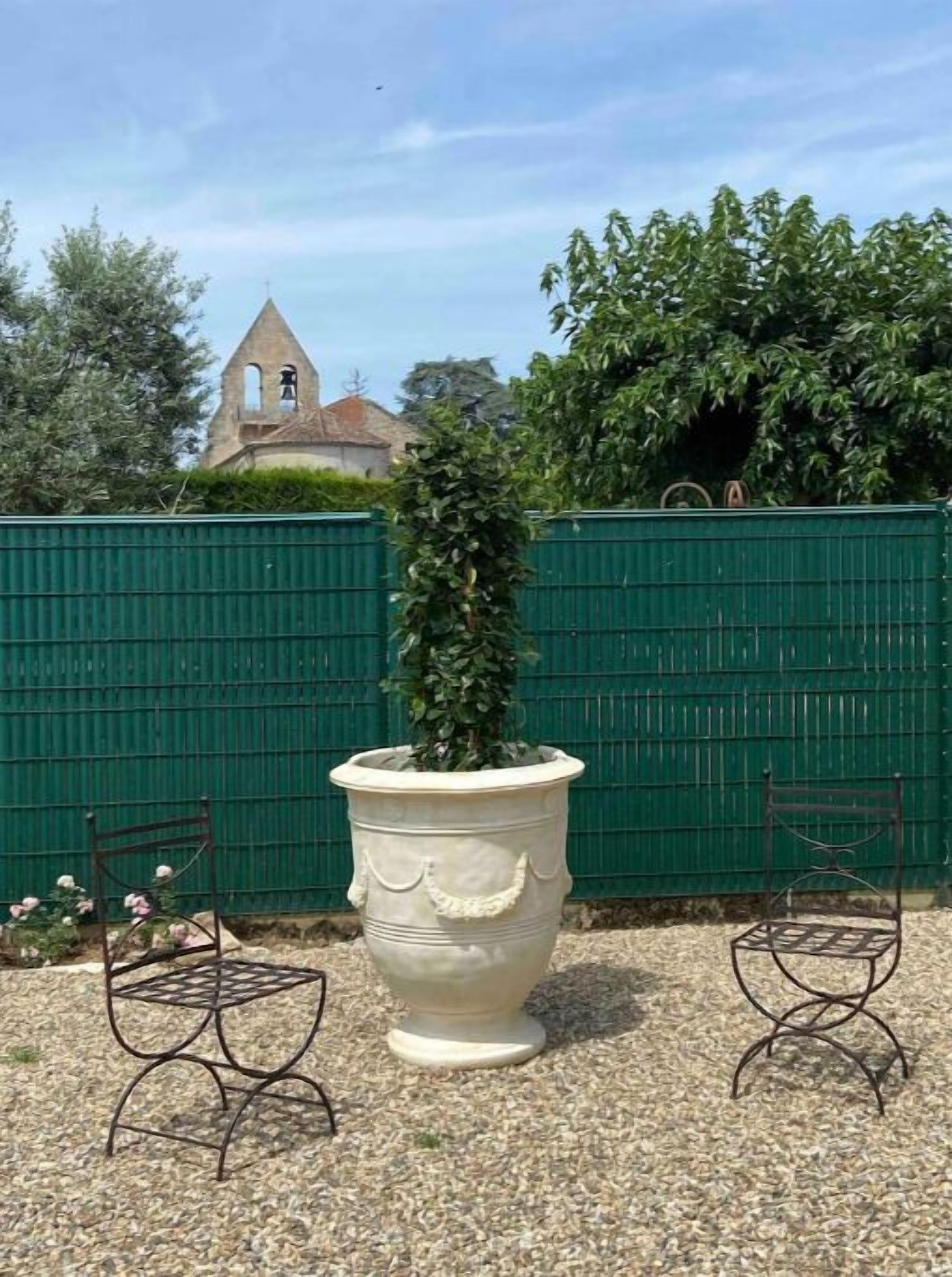 Een grote witte plantenbak met een hoge, slanke plant, twee metalen stoelen en een groene hekken met een kerk op de achtergrond.