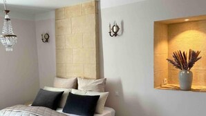 Un dormitorio con cama, una mesita redonda, estante montado en la pared con adornos y una araña.