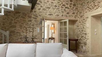 Quarto com paredes de pedra, um sofá branco, uma mesa pequena com abajur, e uma porta que dá para outro cômodo.