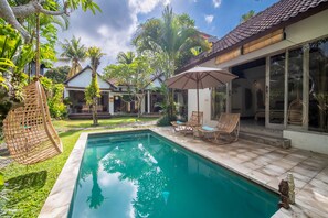 A swimming pool with lounge chairs and an umbrella, surrounded by lush greenery and a house with large windows. - Katak Villa by Villa Finder (Ubud)