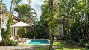 A pool surrounded by lush tropical plants, a wooden deck with chairs, and a stone wall. - Katak Villa by Villa Finder (Ubud)