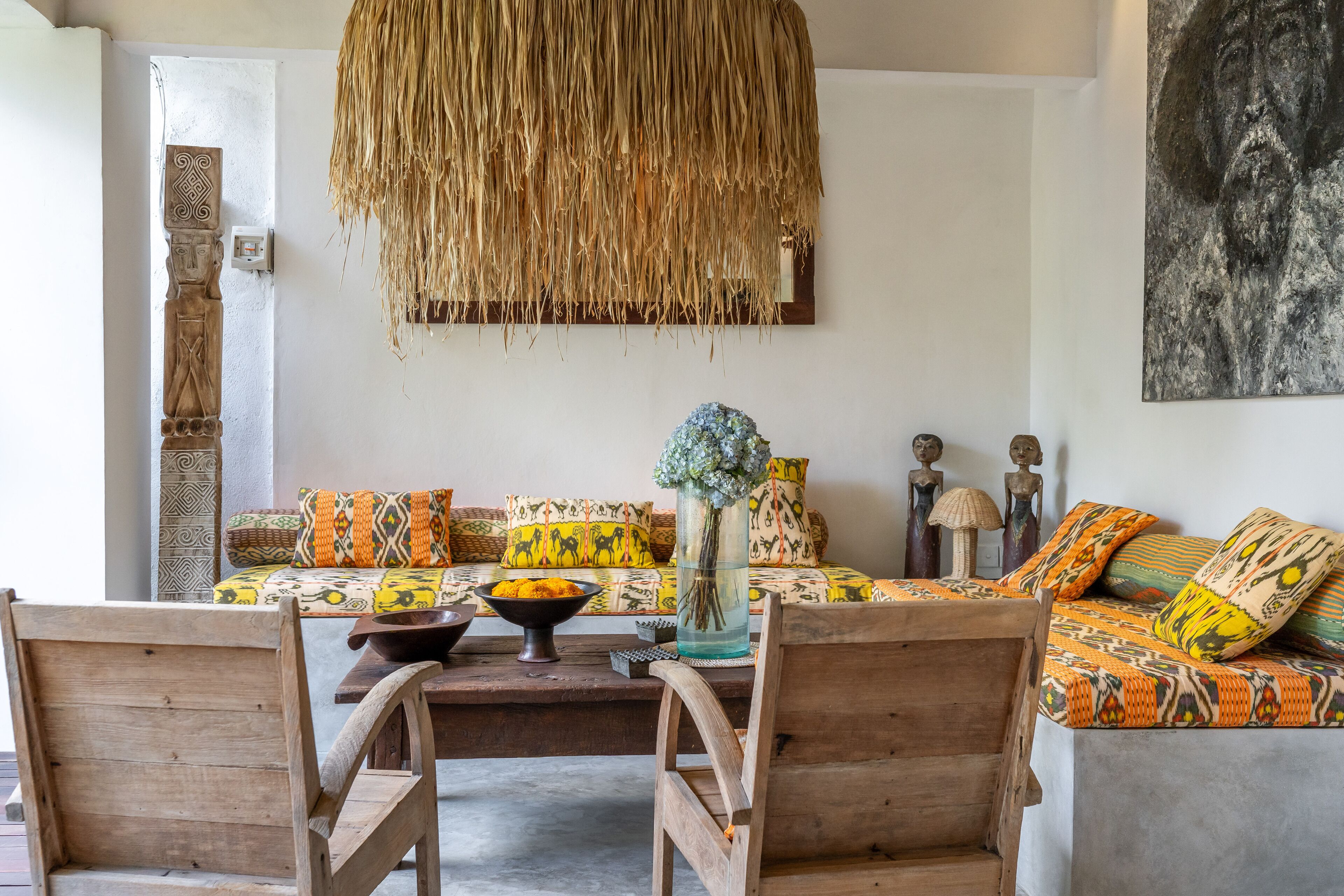 A cozy living room with a sofa set, wooden chairs, a woven hanging light fixture, and a vase with flowers.
