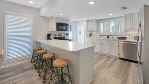 Una cucina moderna con mobili bianchi, un'isola centrale con sgabelli, elettrodomestici in acciaio inossidabile e un'ampia finestra con persiane.