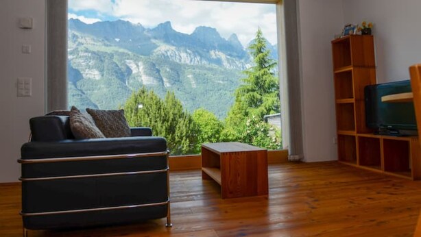A modern living room with a large window offering a mountain view, a dark sofa, a wooden coffee table, and a TV stand. - The Mountains are Calling Retreat (Quarten)