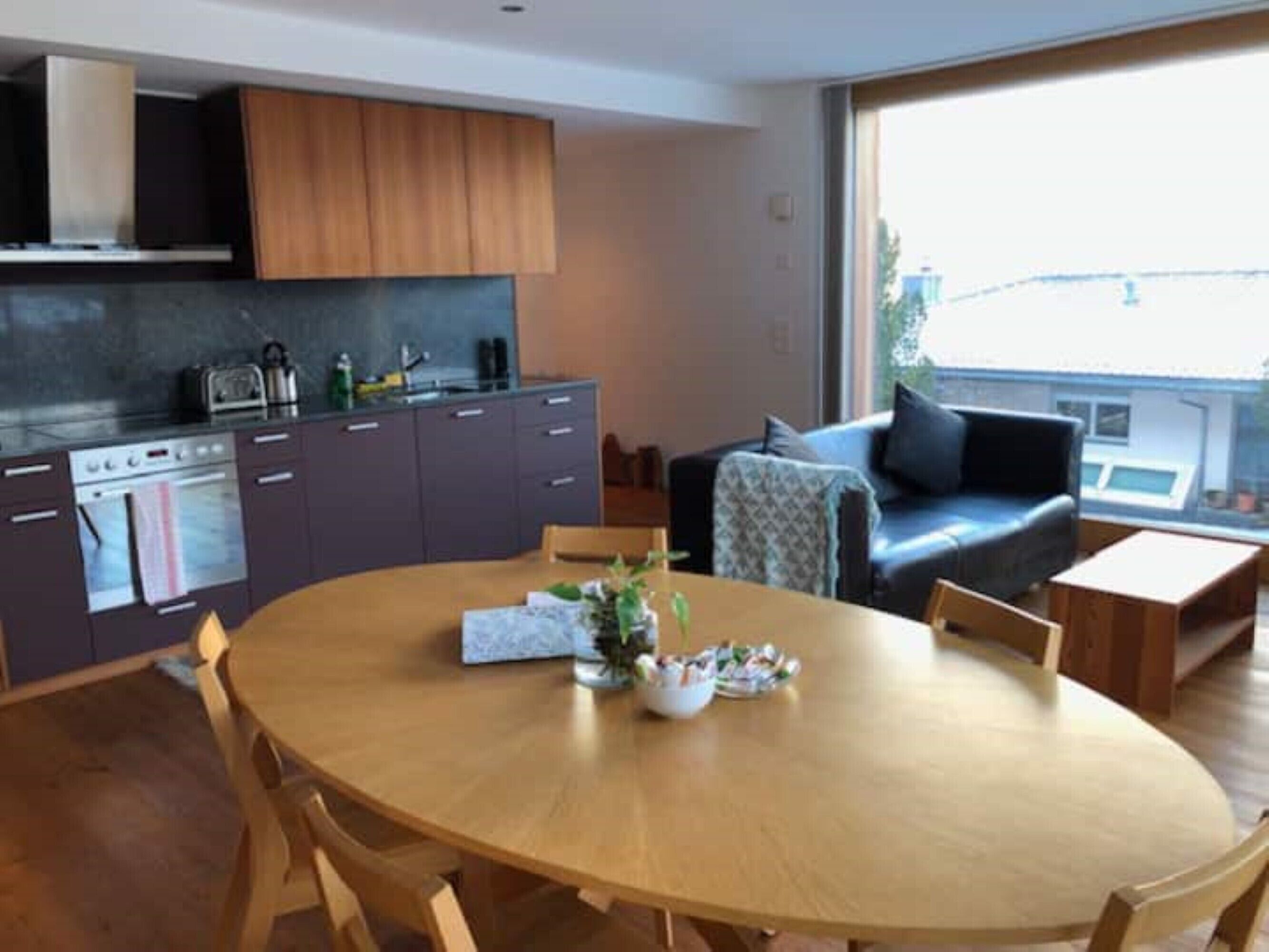 Een moderne keuken met donkere kastjes, een inbouwoven en een eethoek met een ronde houten tafel en stoelen.