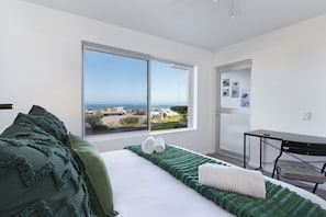 Quarto de hotel moderno com uma grande janela que oferece vista para o mar e prédios, uma cama bem arrumada com roupa de cama verde e branca, e uma pequena escrivaninha com uma cadeira.