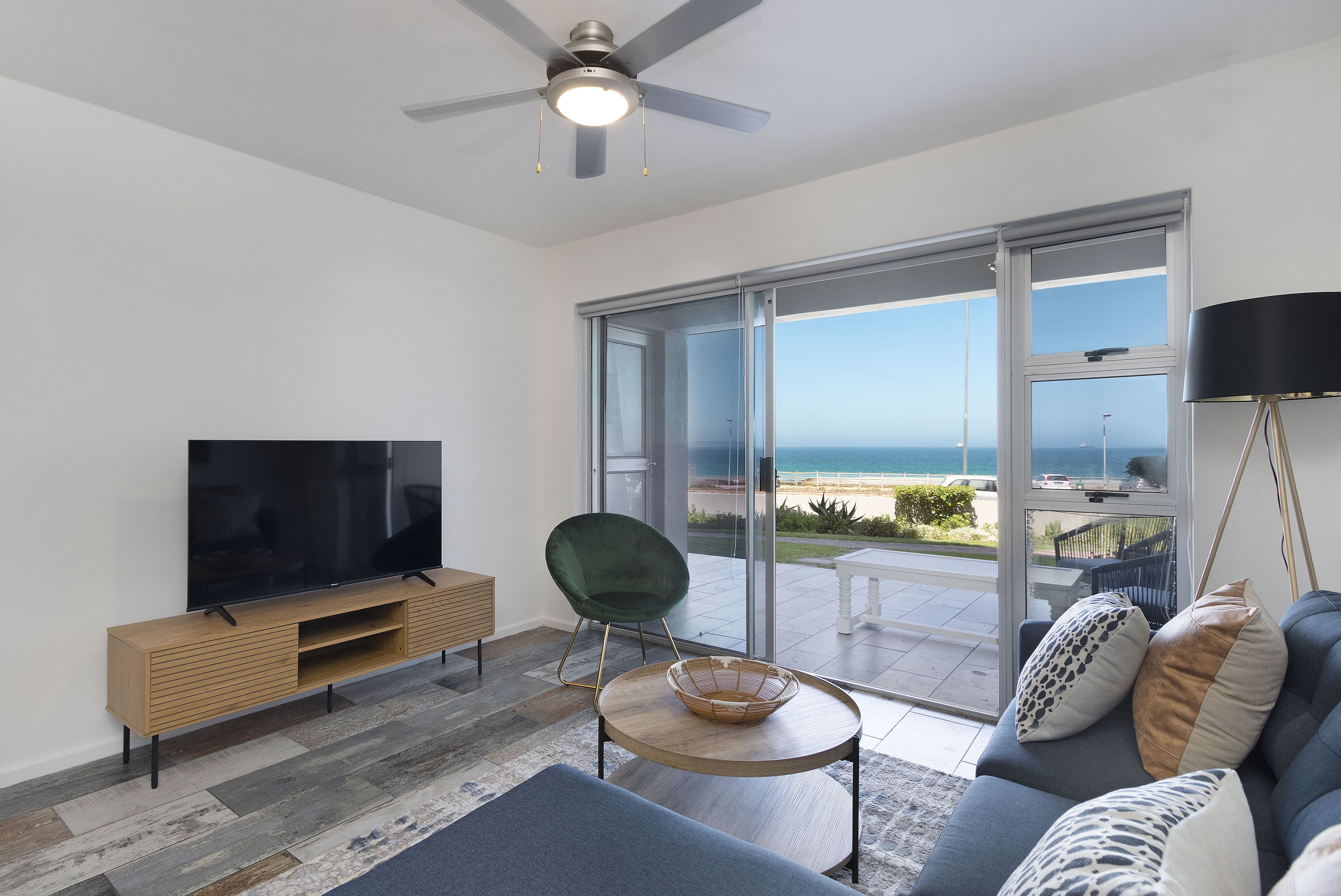 A modern living room with a flat-screen TV, a green chair, a sofa with patterned cushions, a wooden coffee table, and a view of the ocean through large glass doors.