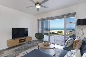 Uma sala de estar moderna com uma TV de tela plana, uma poltrona verde, um sofá com almofadas estampadas, uma mesa de centro de madeira e vista para o oceano através de grandes portas de vidro.