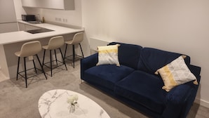A compact living space with a kitchenette, a blue sofa with patterned cushions, a small round table with a vase, and a pair of bar stools. - Captivating 1-bed Apartment in Manchester (Manchester)
