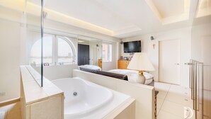 A modern hotel room with a large bathtub, a bed with a white headboard, a bedside table with a lamp, a TV mounted on the wall, and a large window offering a view of the city. - Beautiful 1 Bed on the River (London)