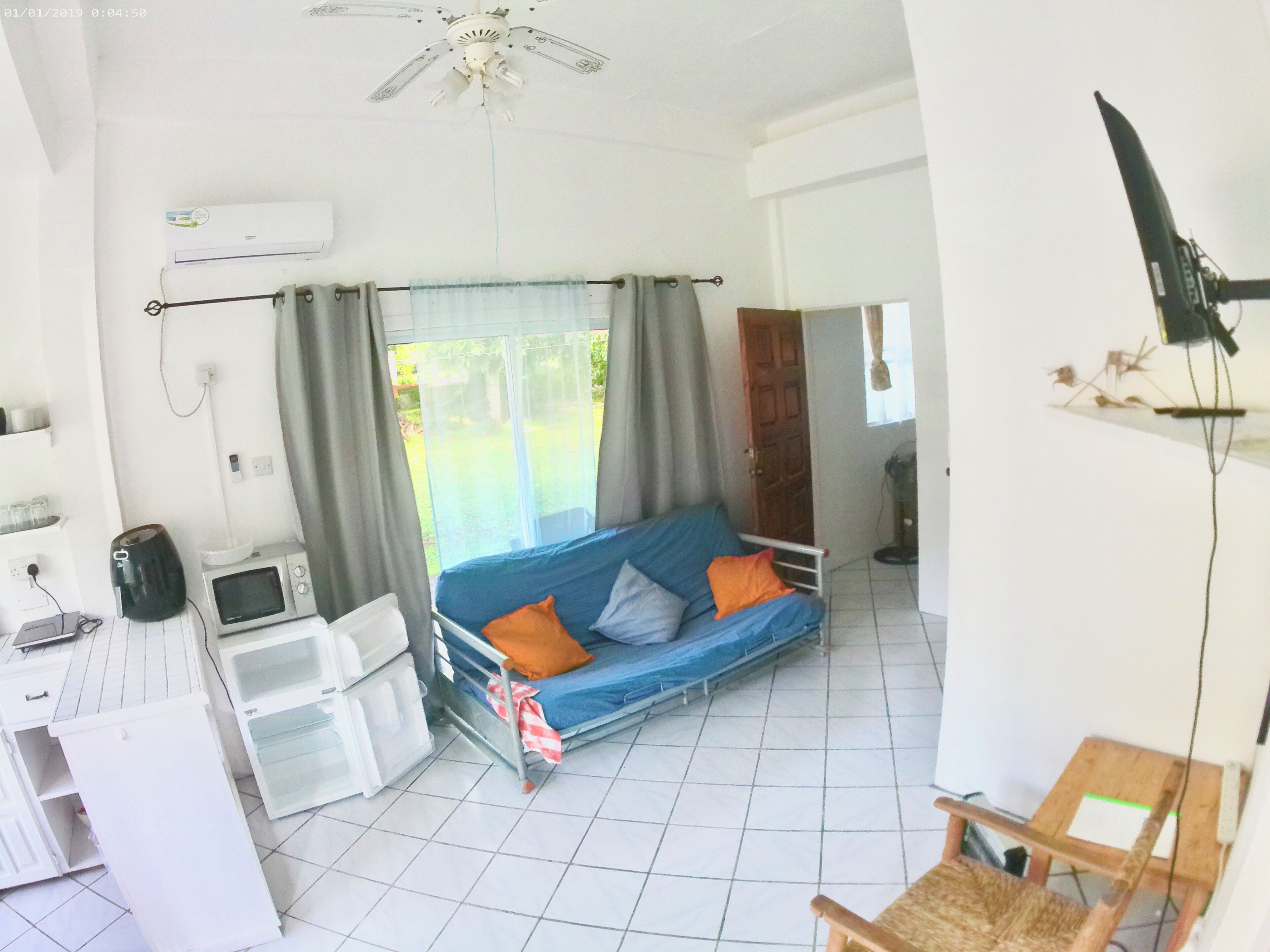 A compact living space with a kitchenette, a blue sofa with orange cushions, a ceiling fan, and a TV mounted on the wall.