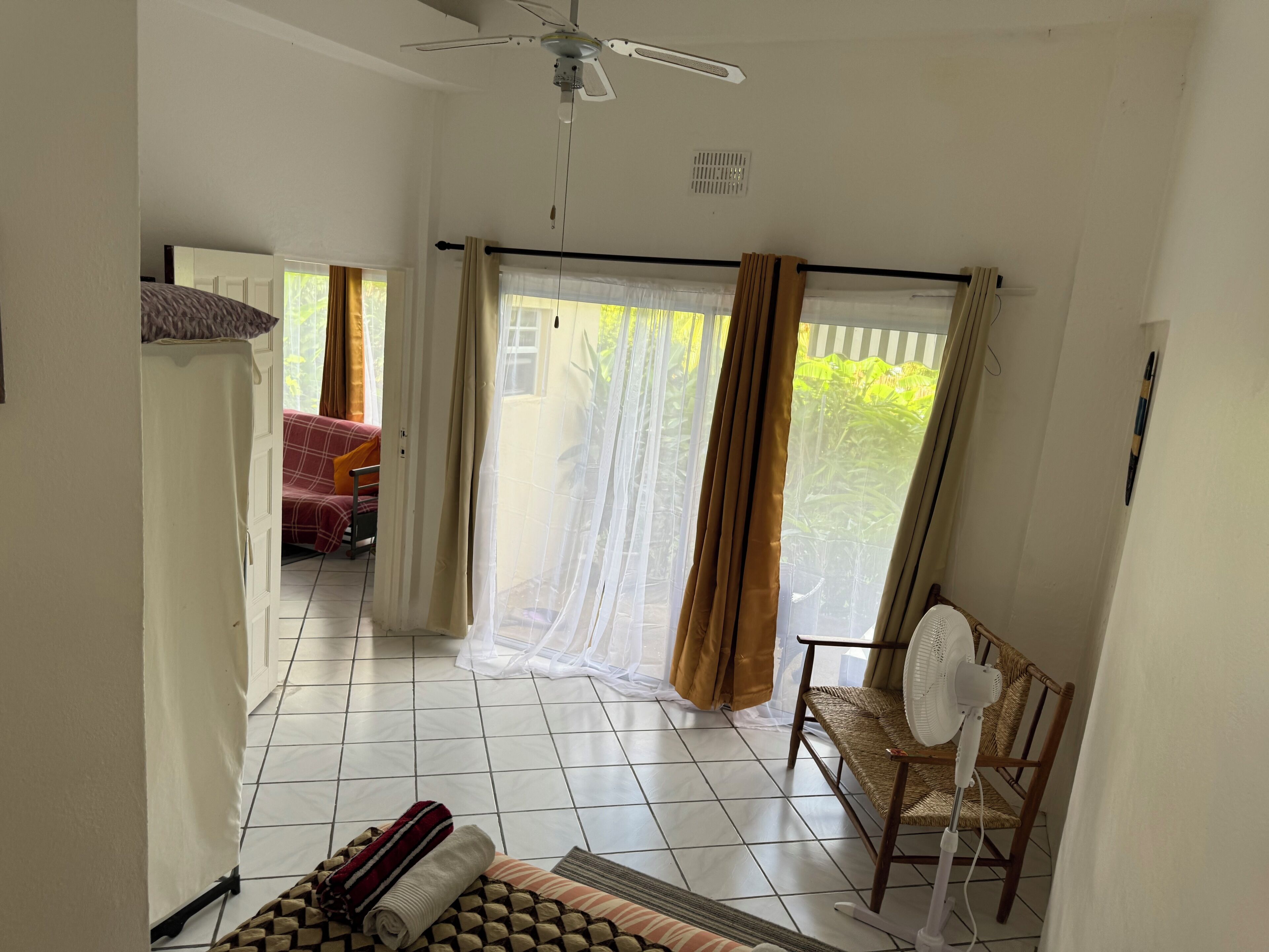 A room with a ceiling fan, a large window with curtains, a wicker chair, and a bed with a red and white striped blanket.