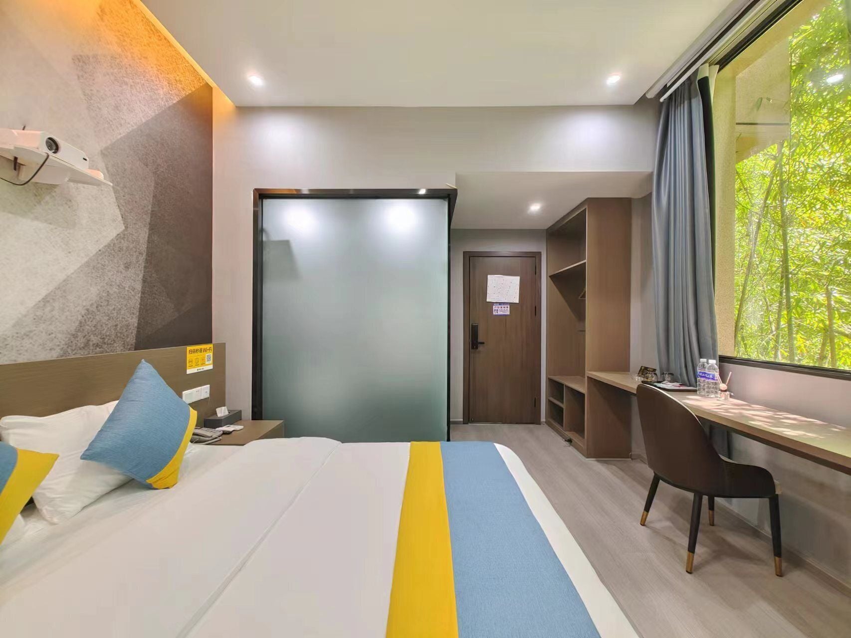 A hotel room with a bed, a desk, a chair, a wardrobe, and a window with a view of trees.