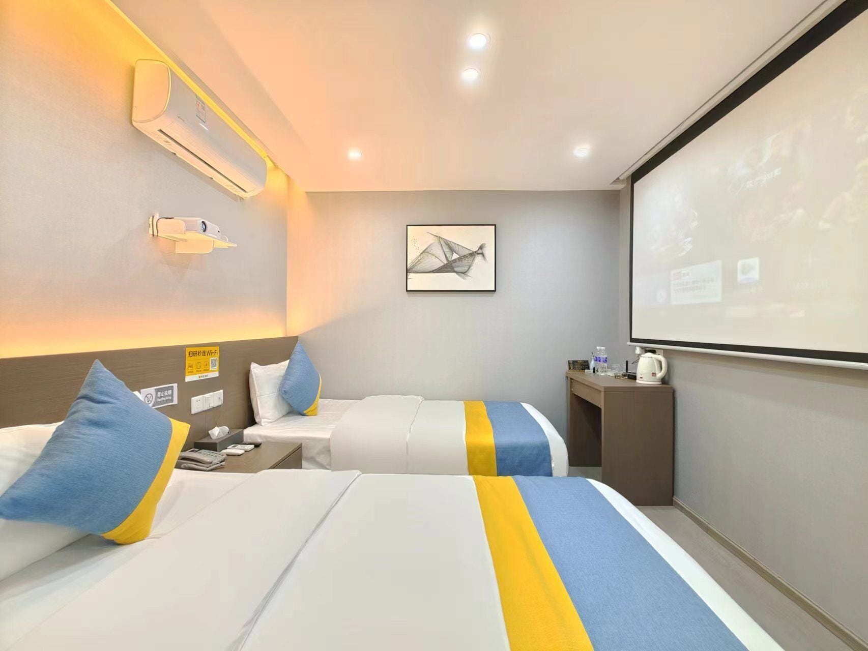 A hotel room with two beds, a desk, a projector screen, and a wall-mounted air conditioner.