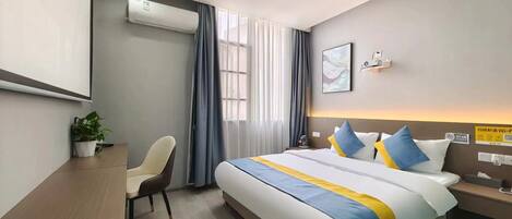 A modern hotel room with a large bed, a desk, and a chair. There is an air conditioning unit on the wall, a window with curtains, and a bedside table with a lamp.
