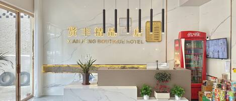 A hotel reception area with a marble reception desk, potted plants, a red vending machine, and a television screen.