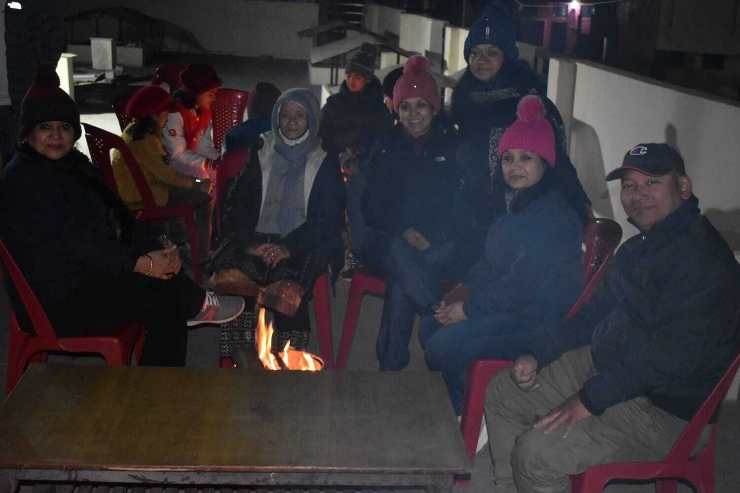 A group of people seated around a fire in an outdoor setting with red plastic chairs and a table.