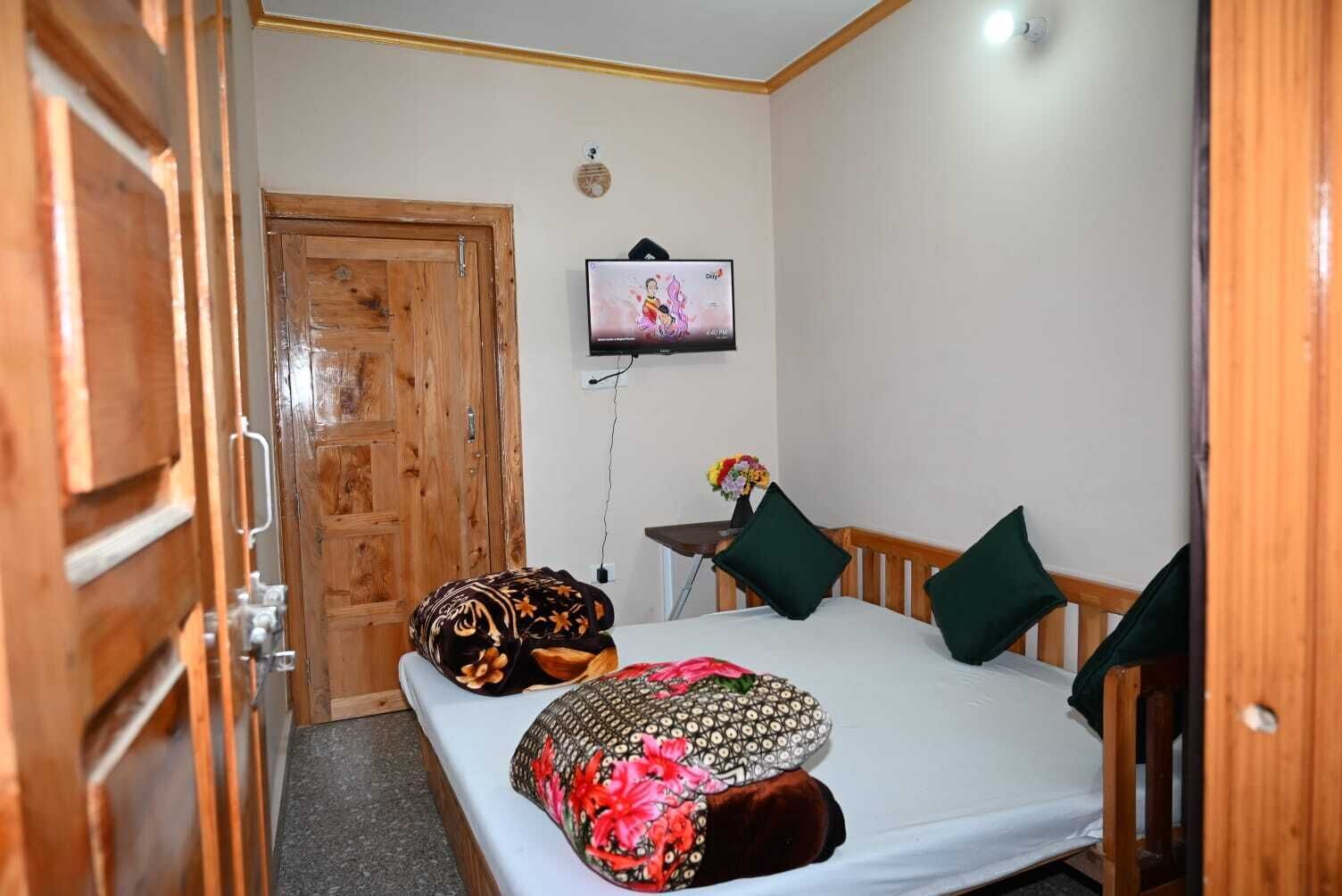 Una habitación pequeña y acogedora, con una puerta de madera, una cama con almohadones estampados, una mesita con un jarrón de flores y un televisor colgado en la pared.