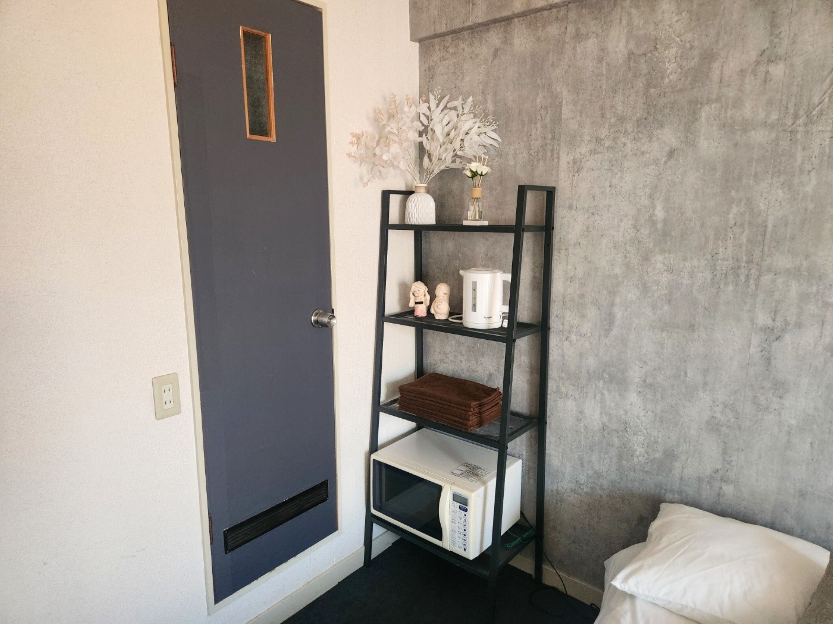 A room with a grey door, a white wall, a concrete wall, a black metal shelf with a white vase, a white kettle, a microwave, and a white pillow.