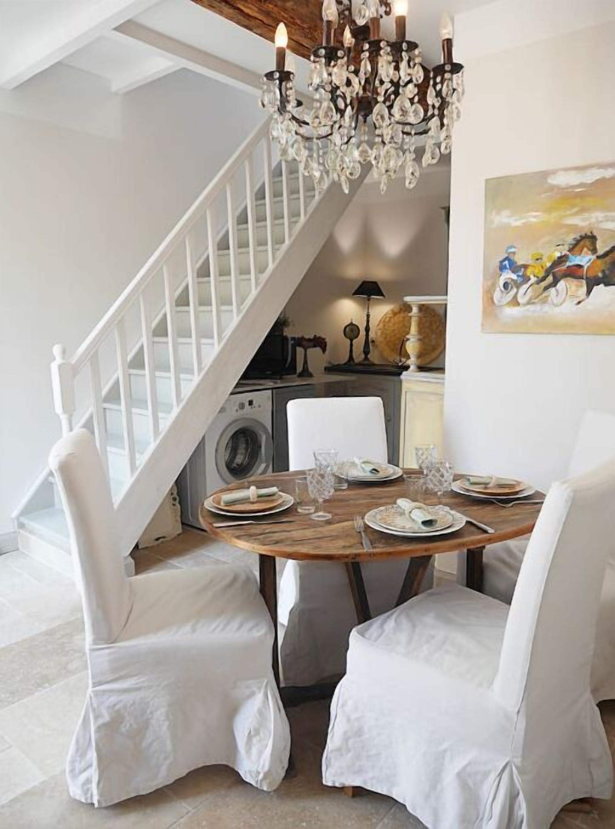 A dining area with a wooden table set for four, white chairs, a chandelier, and a staircase in the background.