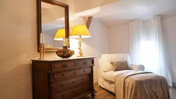 Et soveværelse med en trækommode, et spejl, to lamper, en hvid stol og en seng med et beige tæppe.