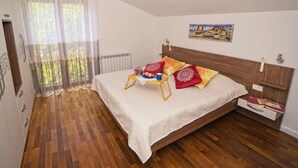 Ein Schlafzimmer mit Holzboden, einem Bett mit weißer Bettdecke, einem kleinen Nachttisch aus Holz und einem Fenster mit transparenten Vorhängen.