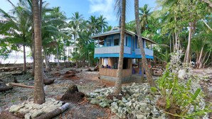 Een blauw, twee verdiepingen tellend huis met balkon, omgeven door palmbomen en een rotsachtig strand.