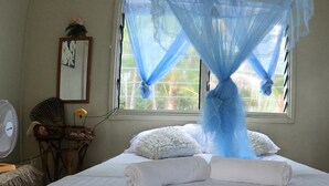 Een netjes opgemaakt bed met witte lakens en een opgerolde handdoek, een nachtkastje met een lamp, een spiegel aan de muur en een raam met een blauw klamboes.