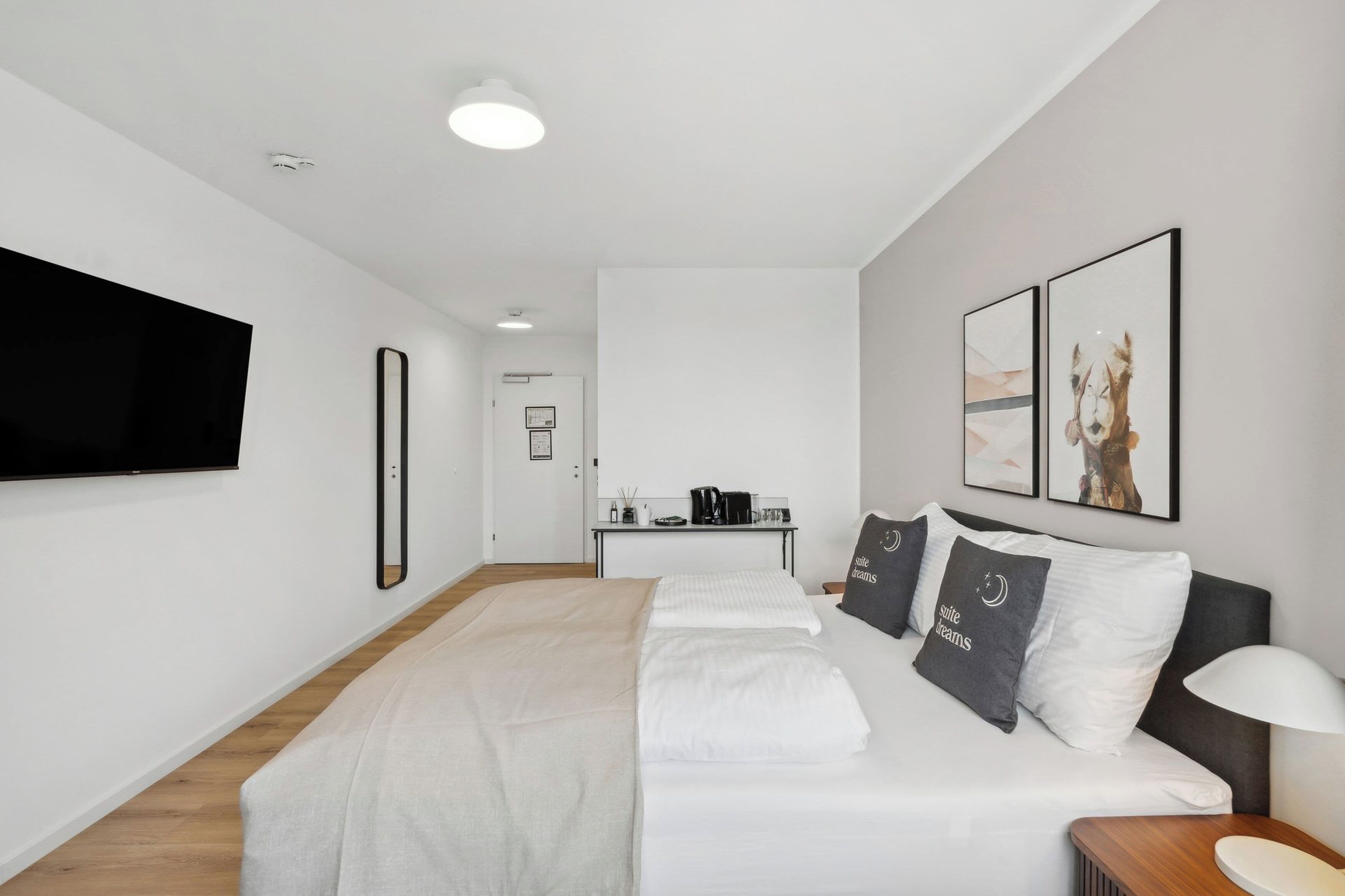 A modern hotel room with a large bed, a flat-screen TV mounted on the wall, and a small table with a lamp.