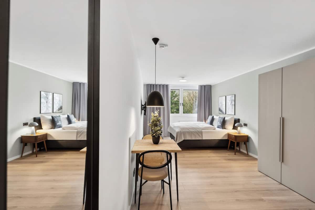 A modern hotel room with two beds, a dining table, and a large window.
