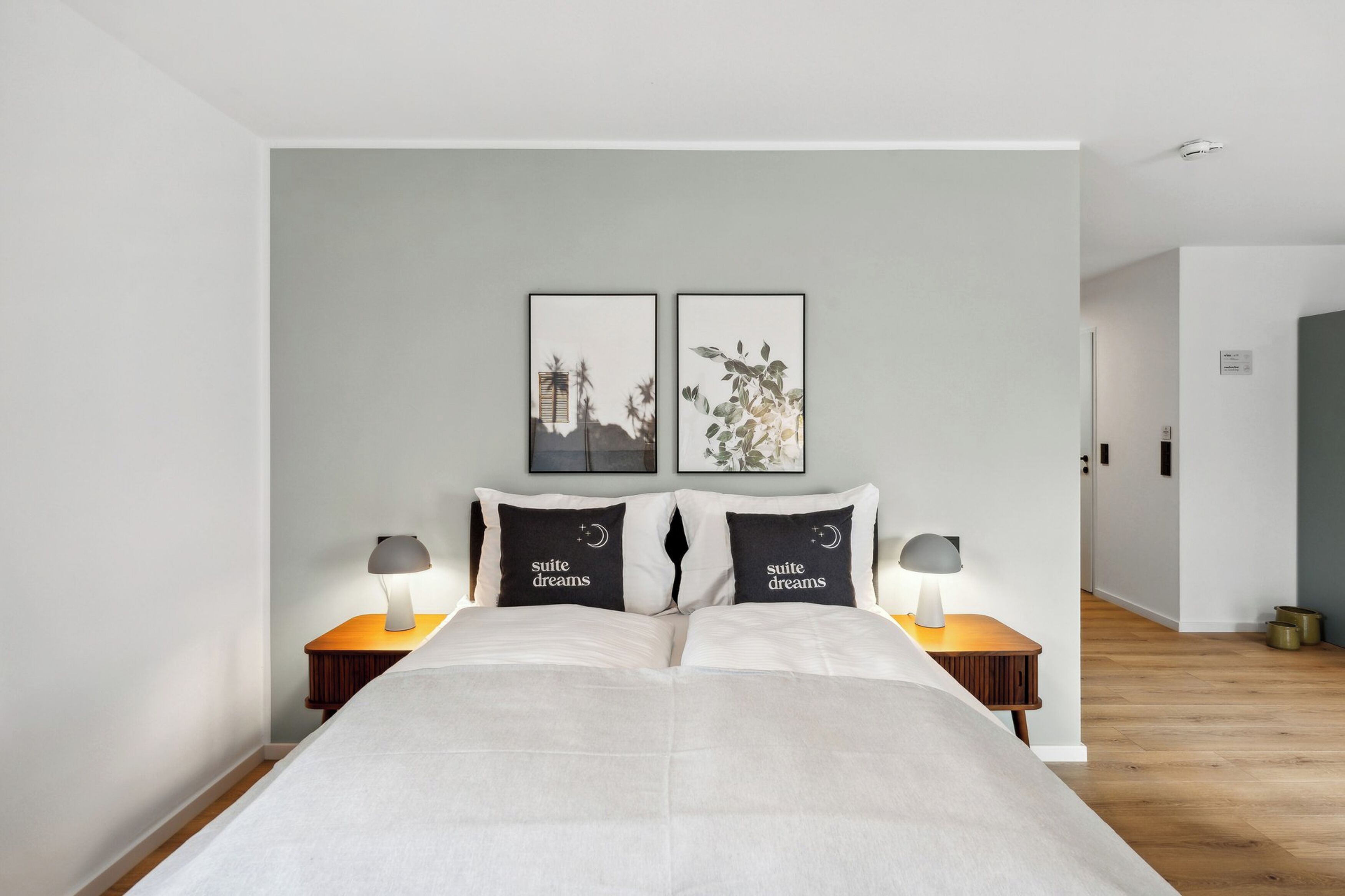 A bedroom with a bed, two bedside tables with lamps, a headboard with two framed pictures, and a hallway leading to another room.