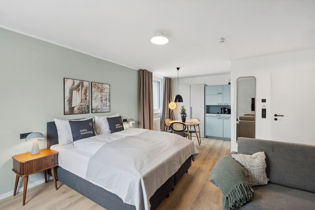 A modern hotel room with a large bed, bedside tables, a sofa, a dining area, and a kitchen in the background.