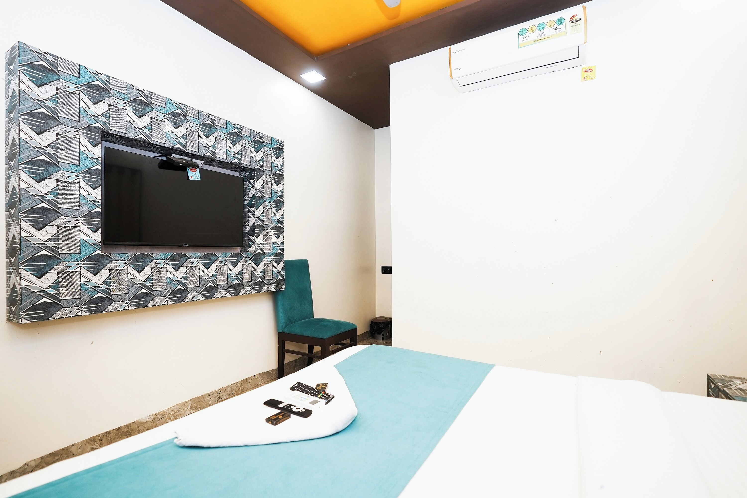 A room with a bed, a TV mounted on a patterned wall, a chair, and an air conditioning unit.
