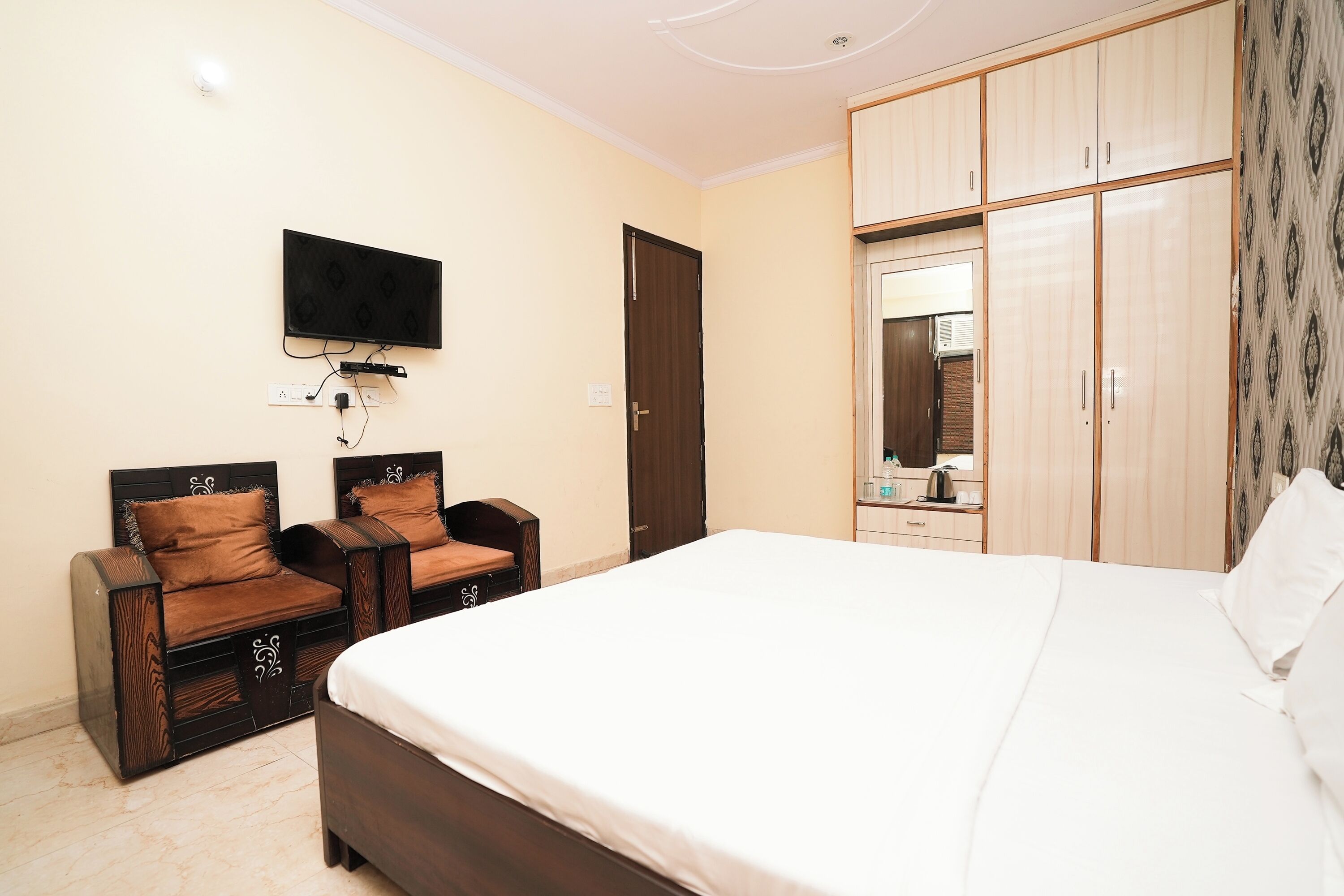 A bedroom with a large bed, two wooden chairs, a television mounted on the wall, and a wardrobe with a mirror.