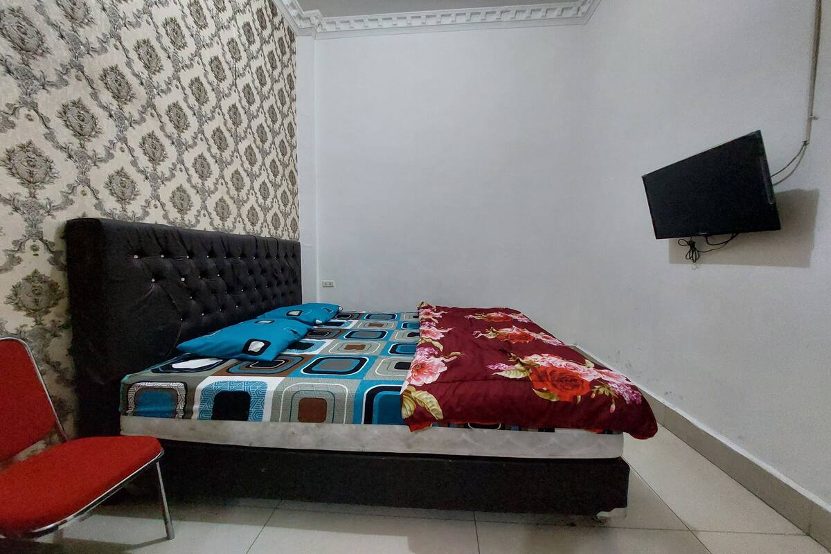 Ein Schlafzimmer mit einem Bett, einem roten Sessel, einem Wandteppich mit Muster und einem an der Wand befestigten Fernseher.