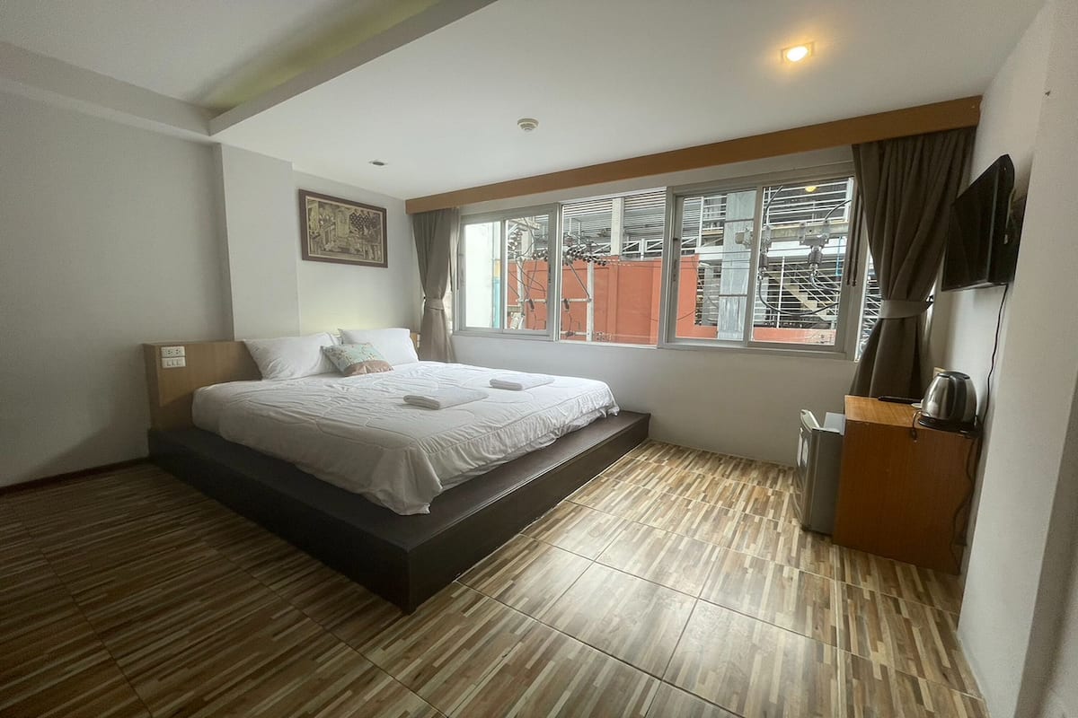 A hotel room with a bed, a small desk with a kettle, a window with curtains, and a television on the wall.