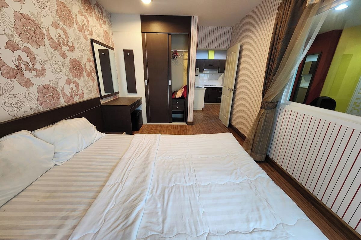 A hotel room with a large bed, a wardrobe, a mirror, and a view of the kitchen.