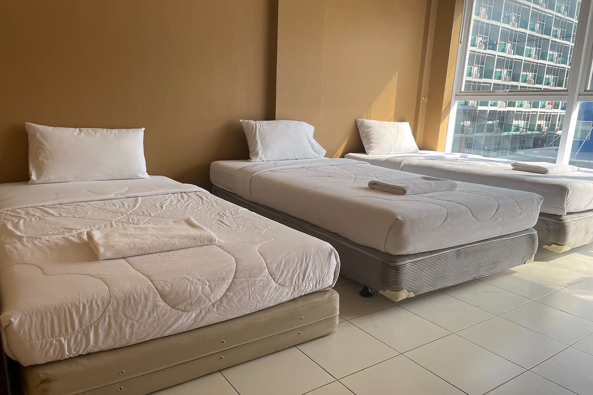 A hotel room with three beds aligned against a wall, a large window with a city view, and a tiled floor.