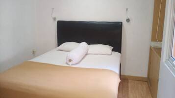 Una cama pequeña y ordenada con cabecera oscura, almohadas blancas y manta color beige.