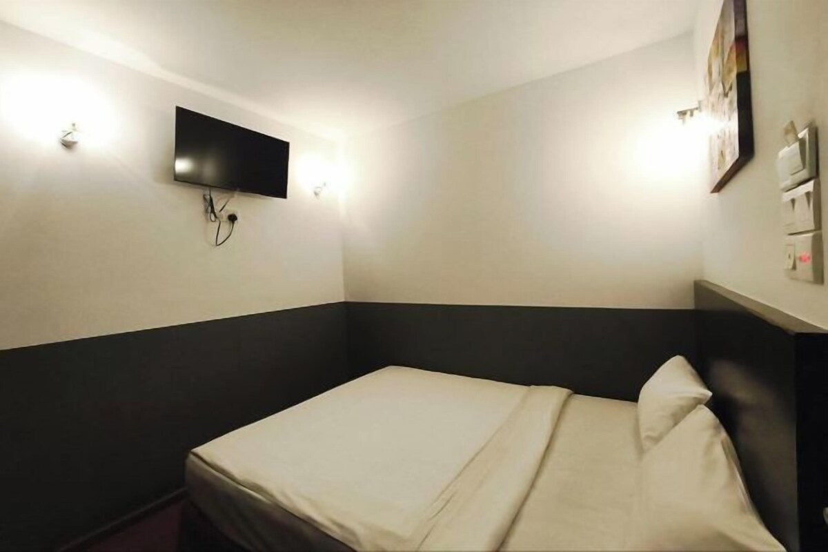 Quarto pequeno com cama de solteiro, parte inferior escura e parte superior clara. Há uma TV de tela plana presa na parede e uma pequena mesa com abajur.