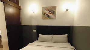 Una cama bien hecha en una habitación de hotel, con cabecera oscura, un cuadro colgado en la pared y dos apliques de luz.