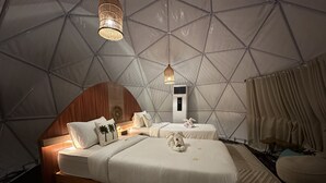 Une pièce confortable, de style tente, bien éclairée, avec deux lits, une tête de lit en bois, une petite table et un luminaire suspendu.