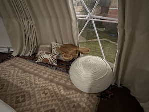 A room with a patterned rug, a round woven pouf, and a wooden side table with a plant. - Pebbles & Fins (Kubu)